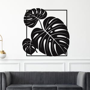 May include: Black and white wall art featuring a stylized depiction of Monstera leaves within a square frame. The leaves have intricate details and are set against a white background, creating a modern, minimalist aesthetic. The artwork is mounted above a gray sofa.