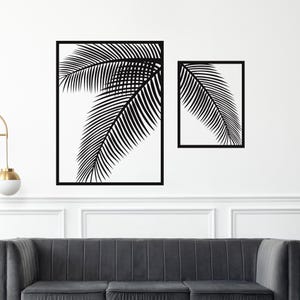 May include: Two black framed wall art pieces featuring palm leaf designs. The larger piece is rectangular, and the smaller piece is square. The palm leaves are black against a white background. The art is mounted on a white wall above a grey sofa.