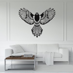 Può includere: Una decorazione murale in bianco e nero raffigurante un uccello con le ali spiegate. Il design dell'uccello presenta dettagliati motivi di piume. L'opera d'arte è montata su una parete bianca sopra un divano bianco.