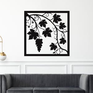 May include: Black metal wall art featuring a grape vine design within a square frame. The vine includes leaves, tendrils, and a cluster of grapes. The art is mounted on a white wall above a gray sofa.