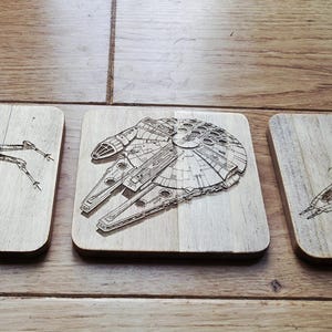Pode incluir: Conjunto de três bases de madeira com imagens gravadas de naves espaciais icónicas de uma popular franquia de ficção científica. Os desenhos incluem um X-wing, a Millennium Falcon e um Y-wing, todos em acabamento de madeira natural.