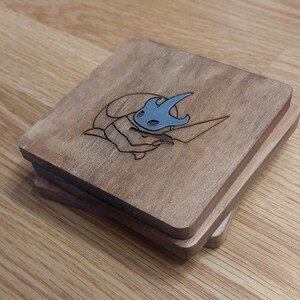 May include: A set of three wooden coasters featuring a laser-etched design of a character from the video game Hollow Knight. The character is a small, blue, insect-like creature with a large head and a pair of wings.