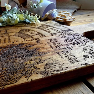 May include: Wooden cutting board with a detailed engraved map design. The board features text including "Moony, Wormtail, Padfoot & Prongs" and other script. The wood has a natural grain and a warm, brown tone.
