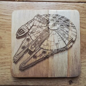 Könnte beinhalten: Ein Holzuntersetzer mit einer detaillierten Lasergravur des Raumschiffs Millennium Falcon. Der Untersetzer ist quadratisch mit abgerundeten Ecken und hat eine hellbraune Oberfläche. Die feinen Details des Raumschiffs sind sichtbar.