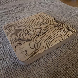 May include: A wooden topographic map of Pen Y Fan, Brecon Beacons, UK. The light wood is etched with dark brown contour lines, showing the terrain. The text "Pen Y Fan" and "Brecon Beacons UK" is visible on the lower left corner.