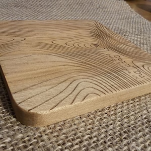 May include: A light brown, square wooden chopping board with a topographic design carved into the surface. The board has rounded corners and the text "Cota Du, Brecon Beacons, UK" is etched on the side.