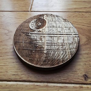 May include: A circular wooden coaster, intricately laser-cut to resemble the Death Star from Star Wars. The coaster is crafted from light brown wood, with detailed surface features. It measures approximately 10 cm in diameter, perfect for protecting surfaces from drink rings.
