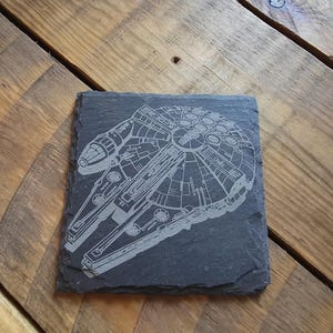 May include: A square, dark grey slate coaster featuring a detailed etched image of the Millennium Falcon starship. The coaster is placed on a wooden surface with a natural grain pattern.