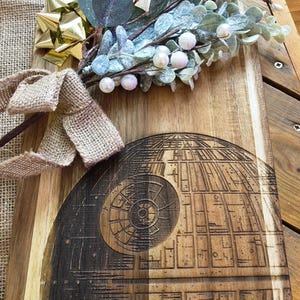 Könnte beinhalten: Holzschneidebrett mit Death Star-Design, mit dunkler, detaillierter Gravur. Das Brett ist mit einer goldenen Schleife, Jutegarn und künstlichem Grün mit weißen Beeren verziert. Das Brett ist aus hellbraunem Holz.