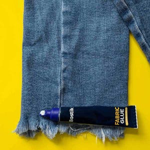 May include: A blue tube of Bostik Fabric Glue on a yellow background. The glue is next to a pair of blue jeans with a frayed hem.