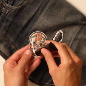 May include: A hand is applying white glue to a colorful sugar skull patch. The patch is being applied to a pair of blue jeans.