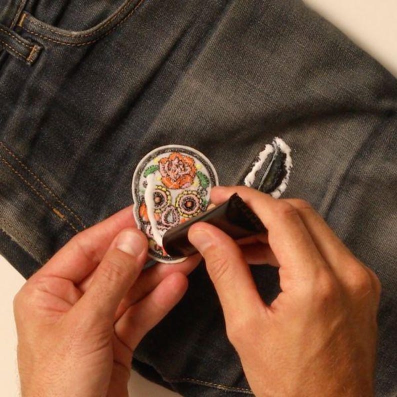 May include: A hand is applying white glue to a colorful sugar skull patch. The patch is being applied to a pair of blue jeans.
