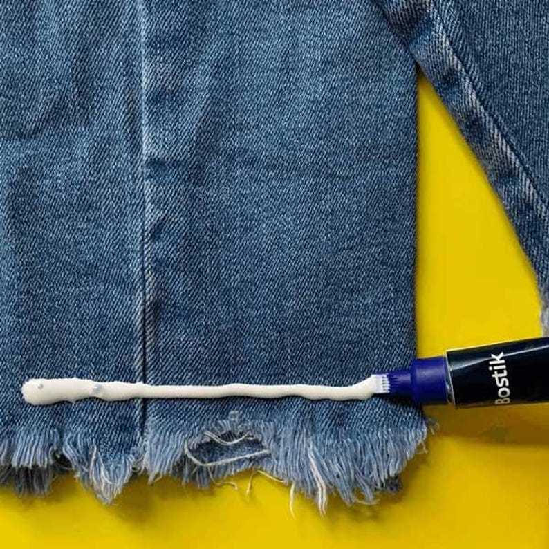 May include: A close-up of a tube of white fabric glue being applied to a ripped pair of blue jeans. The tube is labeled "Bostik".