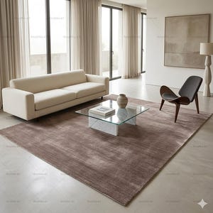 May include: A modern living room with a beige sofa, a glass-top coffee table on a marble base, and a brown rug. A wooden chair with a gray seat and a decorative lamp are also present. Large windows with sheer curtains provide natural light.