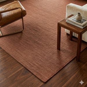 May include: A rectangular, rust-colored rug with a textured surface. The rug is placed on a dark wood floor in a living room setting. A leather chair and a sofa are visible in the background, along with a fireplace.