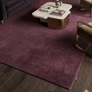 May include: A deep burgundy area rug in a modern living room setting. The rug covers the floor beneath a dark purple velvet sofa, a marble coffee table, and two gold-framed chairs. The room has dark wood flooring.