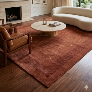 Puede incluir: Salón con una gran alfombra de color óxido con un borde estampado. La habitación incluye un sofá blanco curvo, dos sillones de cuero marrón y una mesa de centro redonda. Obras de arte cuelgan en la pared sobre una chimenea.