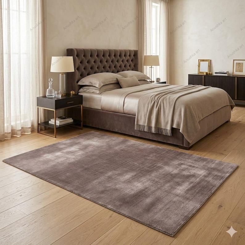 May include: A bedroom scene featuring a large bed with a tufted headboard, neutral-toned bedding, and a matching throw blanket. A large, rectangular, brown rug sits on the wooden floor. Two bedside tables with lamps and a dark dresser complete the room.