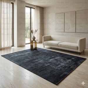 May include: A large, rectangular, dark gray rug dominates the foreground of a living room. The rug has a textured appearance and is placed on a light-colored tiled floor. A beige sofa and a small gold side table with a vase are also visible.
