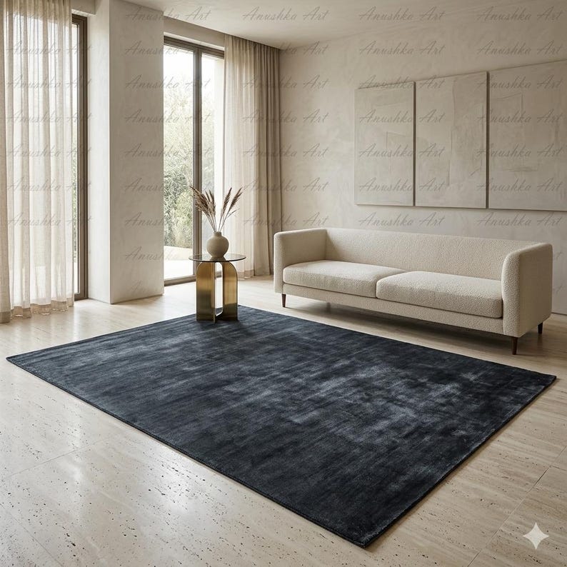 May include: A large, rectangular, dark gray rug dominates the foreground of a living room. The rug has a textured appearance and is placed on a light-colored tiled floor. A beige sofa and a small gold side table with a vase are also visible.