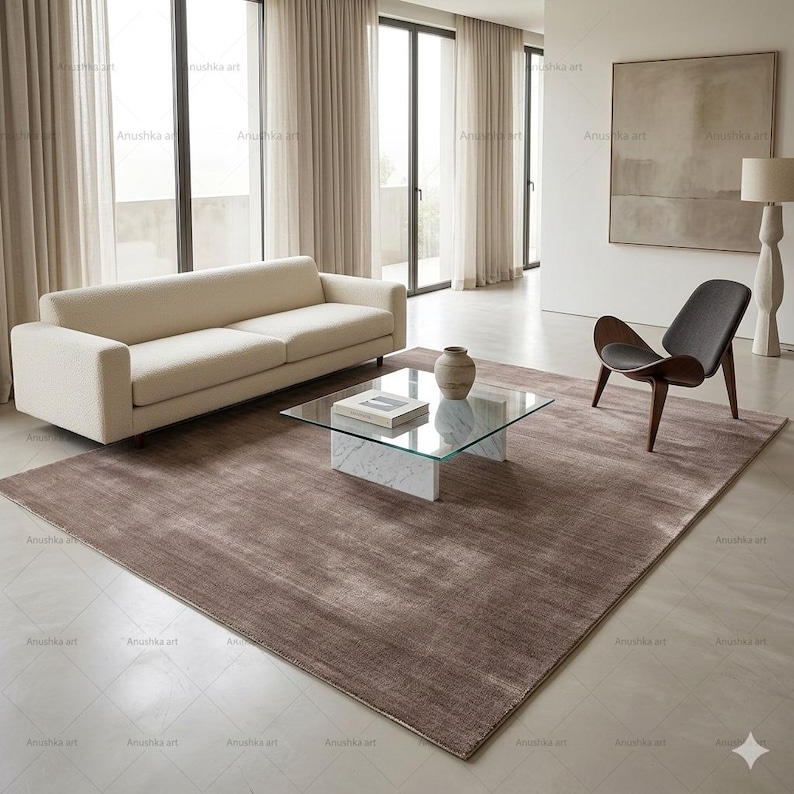 May include: A modern living room with a beige sofa, a glass-top coffee table on a marble base, and a brown rug. A wooden chair with a gray seat and a decorative lamp are also present. Large windows with sheer curtains provide natural light.