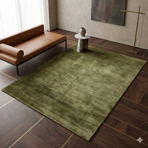 May include: A large, olive green rectangular rug with a soft, textured appearance. The rug is placed on a dark brown, patterned hardwood floor. A brown leather sofa and a small side table are also visible.