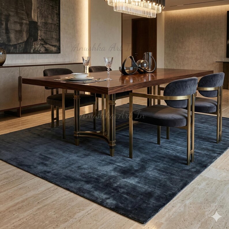 May include: A modern dining room set featuring a dark wood table with brass accents, surrounded by gray velvet chairs. A large blue rug anchors the space, complemented by a chandelier and decorative items on the table.
