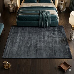 May include: A large, rectangular, dark gray rug with a soft, textured appearance. The rug is placed on a dark wood floor in a bedroom setting, with a teal bed and matching throw blanket visible in the background. The rug adds a touch of luxury to the room.