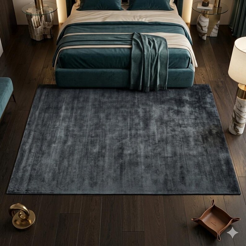 May include: A large, rectangular, dark gray rug with a soft, textured appearance. The rug is placed on a dark wood floor in a bedroom setting, with a teal bed and matching throw blanket visible in the background. The rug adds a touch of luxury to the room.