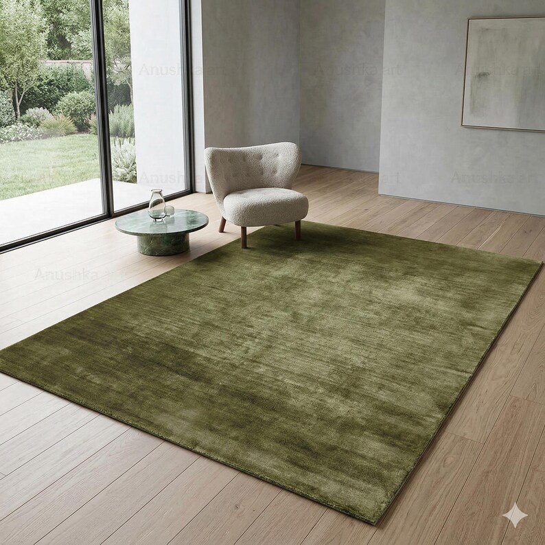 May include: A large, rectangular olive green rug dominates the room. The rug has a textured appearance and covers a significant portion of the light-colored wooden floor. A small, round green marble table and a cream-colored armchair are also visible.