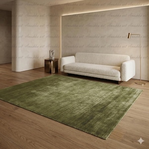 May include: A large, rectangular olive green rug with a textured surface dominates the foreground. A cream-colored sofa and a gold floor lamp are in the background. The room has wooden floors and neutral-toned walls. The text "Anushka art" is repeated across the top.
