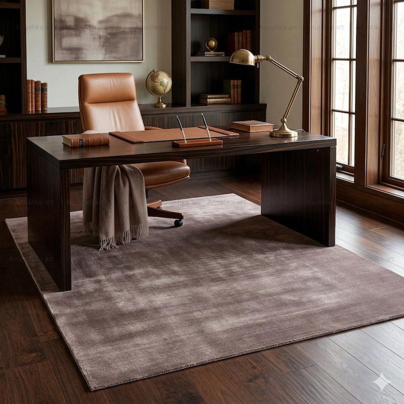 May include: A home office setup featuring a dark wood desk, a brown leather chair, and a matching desk pad. A tan throw blanket is draped over the chair. A rug in shades of gray and brown covers the floor. A brass desk lamp and globe are visible.