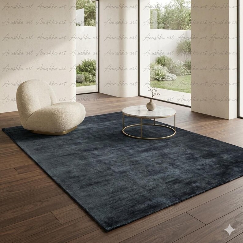 May include: A large, rectangular, dark blue rug dominates a room with a modern aesthetic. A cream-colored armchair and a round marble-topped coffee table with a gold base are also present. The room has large windows and hardwood floors.