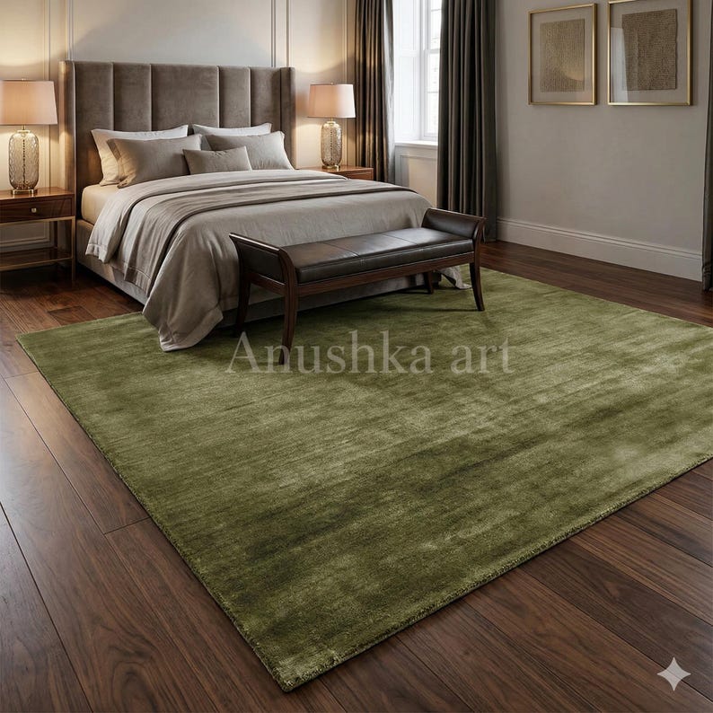 May include: A large, olive green area rug in a bedroom setting. The rug is rectangular with a textured surface, placed on a dark wood floor. The room features a bed with a brown headboard, a bench, and two bedside lamps. The text "Anushka art" is visible.