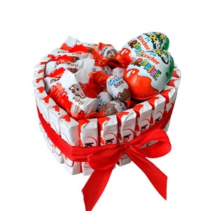Peut inclure: Un panier-cadeau en chocolat en forme de cœur rempli d'œufs Kinder Surprise, de barres chocolatées et d'autres bonbons. Le panier est emballé d'un ruban rouge.
