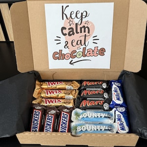Könnte beinhalten: Eine Geschenkbox aus Pappe, gefüllt mit verschiedenen Schokoriegeln, darunter Twix, Mars, Snickers, Bounty und Milky Way. Eine bedruckte Karte in der Box trägt die Aufschrift "Keep calm & eat chocolate".