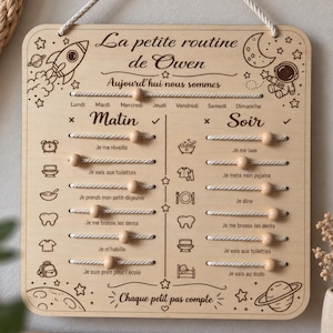 Peut inclure: Un tableau de routine quotidienne en bois clair pour enfant avec des gravures sur le thème de l'espace, incluant une fusée, un astronaute et des planètes. Il comporte des perles en bois coulissantes sur des cordes pour suivre les tâches.
