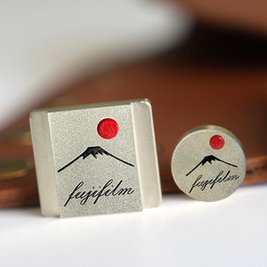 May include: Two silver metal FujiFilm logo charms. The larger charm is rectangular and features a stylized mountain with a red sun. The smaller charm is round and features the same mountain and sun with the FujiFilm logo.