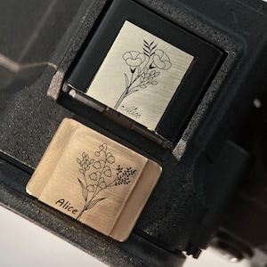 May include: Close-up of a black camera with two custom hot shoe covers. The covers are engraved with floral designs and the name "Alice". One cover is silver, the other is gold.
