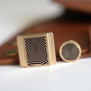 Può includere: Due timbri in metallo dorato con un motivo di illusione ottica in bianco e nero. Il timbro più grande è rettangolare e quello più piccolo è rotondo.