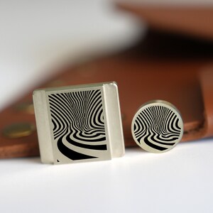 May include: Two silver cufflinks with a black and white swirling pattern. The cufflinks are on a brown leather surface.