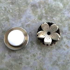 May include: Two silver-toned metal buttons, one with a plain white surface and the other with a detailed cherry blossom design.