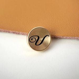 Op de afbeelding: Een kleine, ronde, goudkleurige metalen knoop met een zwarte, cursieve letter "V" in het midden. De knoop is geplaatst tegen een achtergrond van bruin leer en een wit oppervlak.