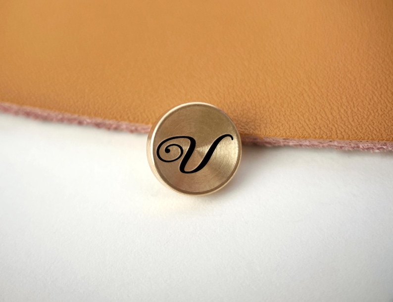Op de afbeelding: Een kleine, ronde, goudkleurige metalen knoop met een zwarte, cursieve letter "V" in het midden. De knoop is geplaatst tegen een achtergrond van bruin leer en een wit oppervlak.