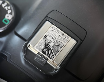 May include: A silver lighter featuring a black and white engraving of Edvard Munch's "The Scream" artwork. The lighter is rectangular with rounded edges and is set against a black background.