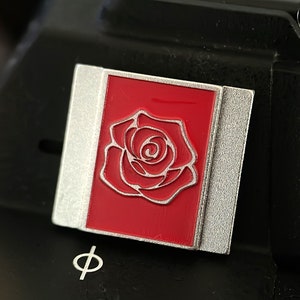May include: A silver and red enamel pin featuring a white outline of a rose.