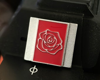 May include: A silver and red enamel pin featuring a white outline of a rose.