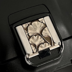 May include: A close-up of a black camera with a silver flash attachment. The flash attachment has a decorative design of ginkgo leaves.