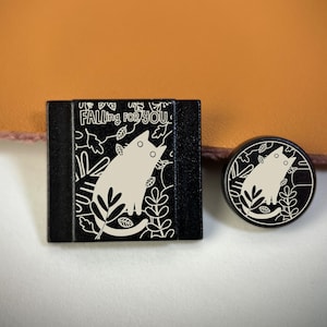May include: Black and white cat-themed accessories. A square, black object with the text "Falling for You" and a white cat illustration. A round, black object with a similar cat design.