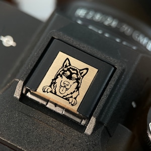 May include: A black camera with a gold button featuring a black and white illustration of a husky dog with its tongue sticking out.